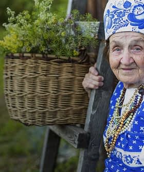Elderly woman wearing traditional blue and white attire with a floral headscarf, holding a wooden post beside a basket of flowers. - Olive Oil Times