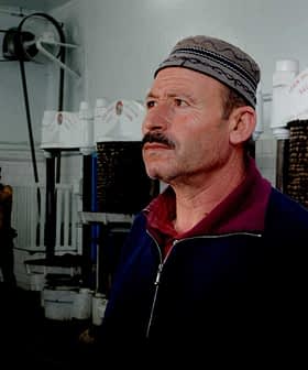 Man wearing a traditional hat standing inside an oil pressing facility with machinery in the background. - Olive Oil Times