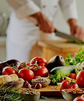 A variety of fresh vegetables, herbs, and cooking ingredients arranged on a kitchen counter with a chef preparing food in the background. - Olive Oil Times