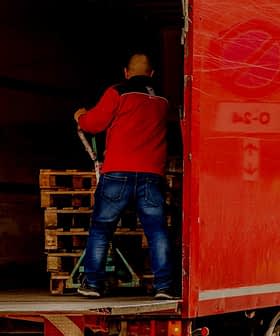 A man in a red jacket loading wooden pallets into the back of a red delivery truck. - Olive Oil Times