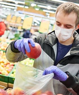 Man in a mask and gloves placing an apple into a plastic bag in a grocery store. - Olive Oil Times
