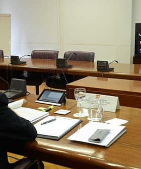 Man attending a virtual meeting with multiple participants displayed on a screen in a conference room. - Olive Oil Times