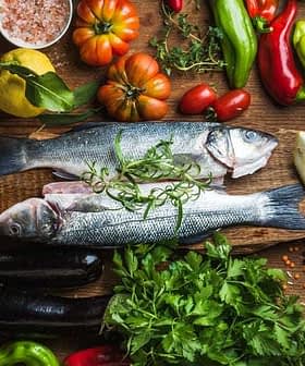 Two whole fish placed on a wooden board surrounded by various vegetables and herbs. - Olive Oil Times