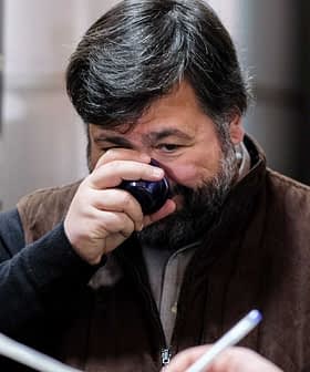 Man smelling olive oil from a small cup while another person observes in the background. - Olive Oil Times