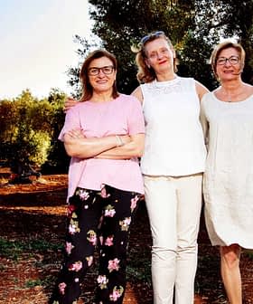 Three women standing together in an olive grove, wearing casual clothing and smiling at the camera. - Olive Oil Times