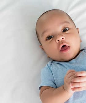 A baby with short hair and a blue shirt lying on a white surface, looking directly at the camera. - Olive Oil Times