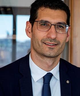 Man wearing glasses and a suit, seated in front of a wooden cabinet with books and decorative items. - Olive Oil Times