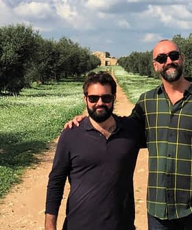Two men posing together in an olive grove with trees and grass in the background. - Olive Oil Times