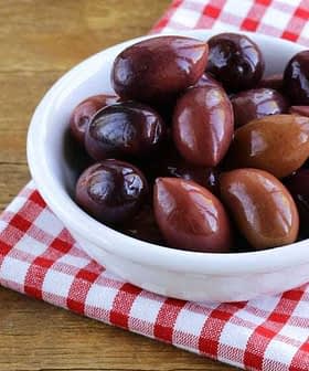 A bowl filled with various types of olives on a red and white checkered cloth. - Olive Oil Times