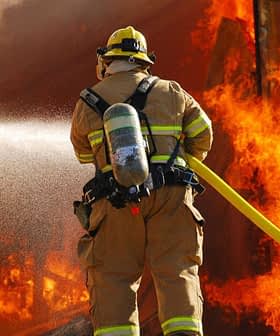 Firefighter in protective gear using a hose to extinguish flames from a burning structure. - Olive Oil Times