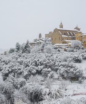 A snow-covered village with buildings and trees blanketed in white snow on a hillside. - Olive Oil Times