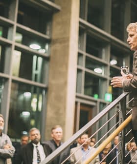 A man in a blazer speaking to an audience while holding a glass in a well-lit indoor space. - Olive Oil Times