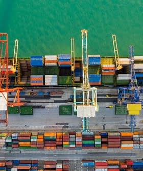 Aerial view of a cargo ship docked at a port with cranes and stacked containers. - Olive Oil Times