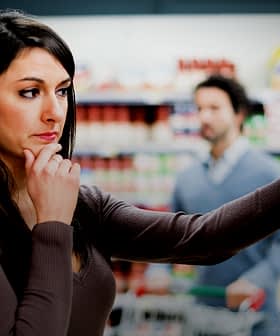 Woman with long dark hair contemplating products in a grocery store aisle while a man is in the background. - Olive Oil Times