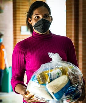 A woman wearing a pink sweater and black mask holds a bag of food items in a corridor. - Olive Oil Times