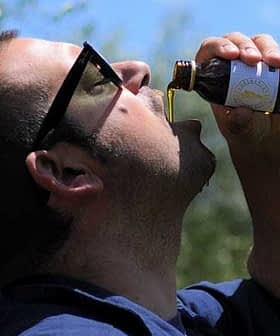 Man tilting his head back while drinking olive oil from a small bottle outdoors. - Olive Oil Times