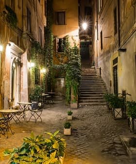 Narrow street featuring outdoor tables and potted plants, illuminated by street lamps at night. - Olive Oil Times