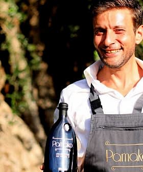 Man in an apron holding two bottles of Pamako olive oil outdoors with trees in the background. - Olive Oil Times