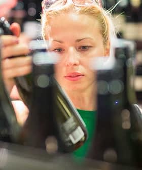 Woman examining a wine bottle while shopping in a store with multiple bottles in the background. - Olive Oil Times