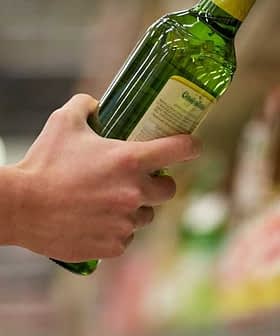A person holding a green bottle of olive oil while reaching for another on a shelf. - Olive Oil Times
