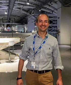 A man wearing a checked shirt and beige pants standing in a modern indoor area with a bar-like setup. - Olive Oil Times
