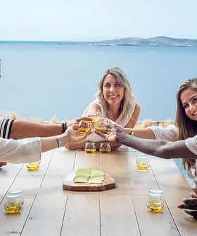 Five individuals sitting at a table, raising glasses in a toast with a scenic view of water in the background. - Olive Oil Times