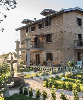 Two-story stone house with balconies and a garden featuring trees and shrubs. - Olive Oil Times
