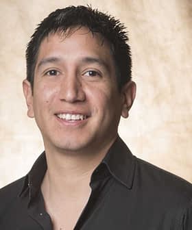 A man with short black hair wearing a black shirt, smiling at the camera against a neutral background. - Olive Oil Times
