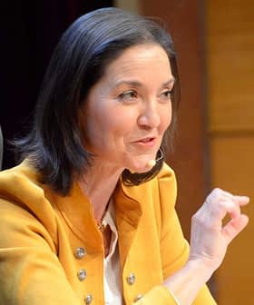 A woman in a yellow jacket speaking at a conference with a microphone and a water bottle on the table. - Olive Oil Times
