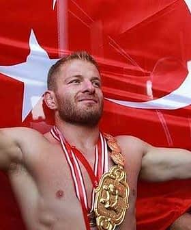 A wrestler celebrating with arms outstretched in front of a large Turkish flag, wearing a championship belt and medals. - Olive Oil Times