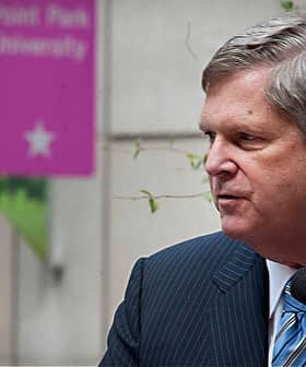 A man in a striped suit speaking into a microphone at a public event with signage in the background. - Olive Oil Times