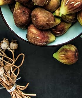 A bowl filled with fresh figs alongside a bundle of dried poppy seed pods on a dark surface. - Olive Oil Times