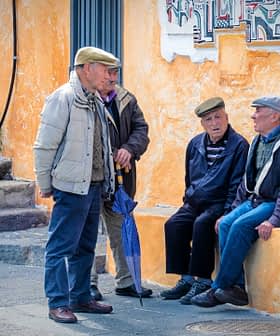 Four elderly men wearing caps engaged in conversation while seated and standing near a colorful wall. - Olive Oil Times