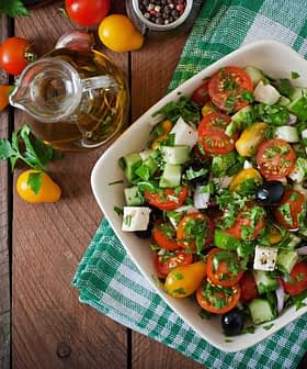 A bowl of fresh vegetable salad with tomatoes, cucumbers, olives, and herbs on a wooden table. - Olive Oil Times