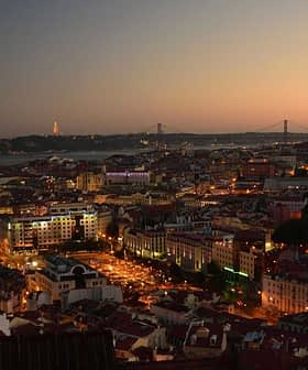 A panoramic view of Lisbon cityscape at dusk with lights illuminating the buildings and the bridge in the background. - Olive Oil Times