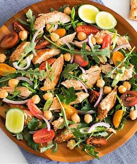 A large wooden bowl filled with a mixed salad featuring grilled chicken, arugula, tomatoes, and chickpeas. - Olive Oil Times