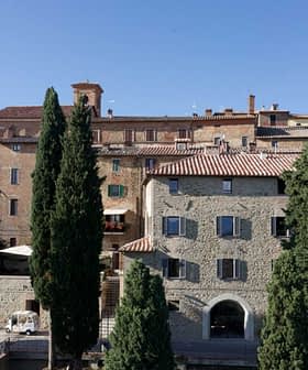 Stone buildings with terracotta roofs in a Tuscan village, surrounded by tall trees. - Olive Oil Times