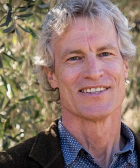 Man with gray hair and a blue shirt smiling in front of olive trees. - Olive Oil Times