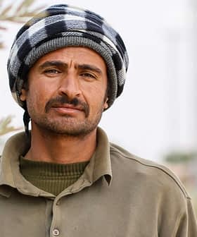 A man wearing a traditional black and white checked headscarf and a green shirt, standing outdoors. - Olive Oil Times