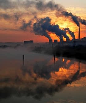 Silhouetted industrial power plant with smoke stacks against a sunset sky and water reflection. - Olive Oil Times