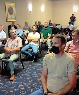 Group of people seated in a meeting room, some wearing masks, listening to a speaker. - Olive Oil Times