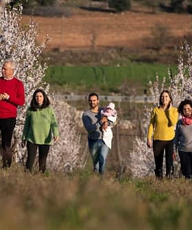 Group of six people walking through a blooming orchard with white flowers on trees. - Olive Oil Times