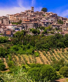 A hilltop village with stone buildings and olive groves in the foreground under a blue sky. - Olive Oil Times