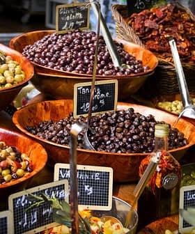 Assorted olives displayed in wooden bowls at a market, with labels indicating prices. - Olive Oil Times
