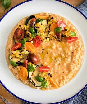 Bowl of creamy risotto topped with grilled vegetables and cherry tomatoes on a wooden table. - Olive Oil Times