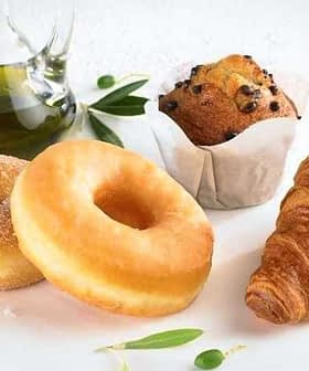 Two sugar-coated donuts, a chocolate chip muffin, and a croissant on a plate with a glass of olive oil. - Olive Oil Times