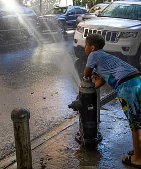 A child in swim trunks leaning against a fire hydrant with water spraying out onto the street. - Olive Oil Times