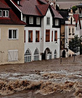 Residential buildings partially submerged in floodwaters with strong currents visible. - Olive Oil Times