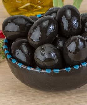 A bowl filled with black olives on a wooden surface with herbs and spices nearby. - Olive Oil Times
