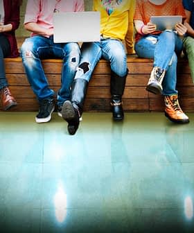 A group of individuals sitting on a bench, each using various electronic devices such as laptops and tablets. - Olive Oil Times